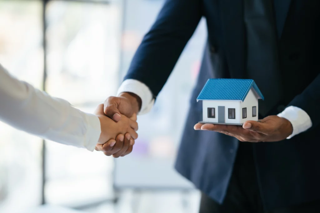 a businessman holding a house model on one hand while shaking a businesswoman's hand.
